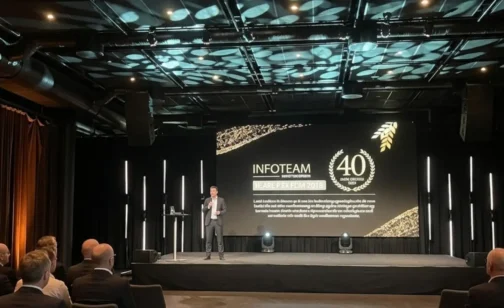 Speaker presenting on stage at an Infoteam event, with a large screen showing ‘Infoteam 40 years’ anniversary branding and an audience seated in front; Novacura was a sponsor.