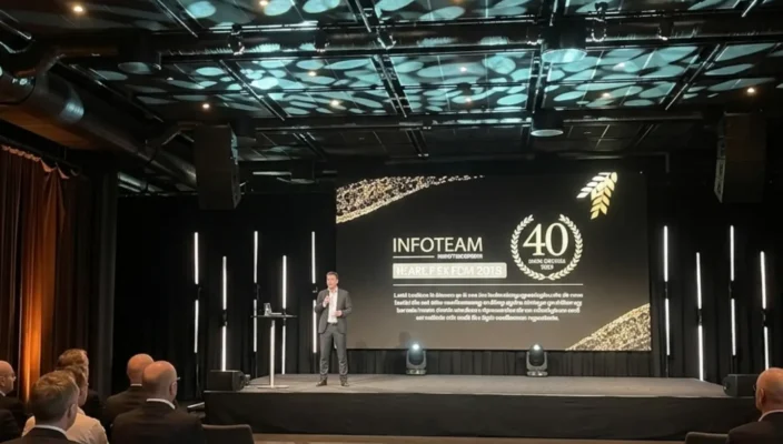 Speaker presenting on stage at an Infoteam event, with a large screen showing ‘Infoteam 40 years’ anniversary branding and an audience seated in front; Novacura was a sponsor.