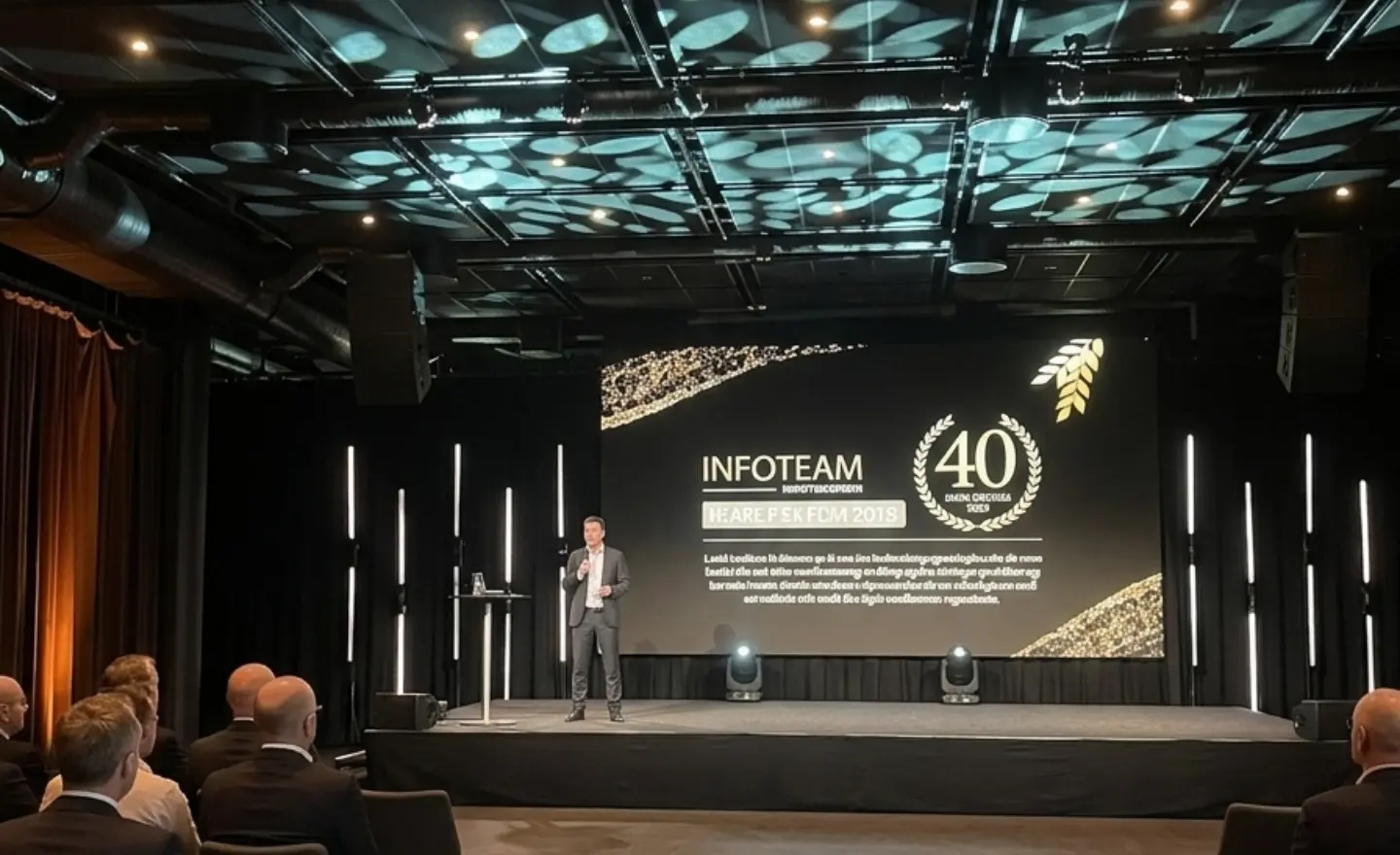 Speaker presenting on stage at an Infoteam event, with a large screen showing ‘Infoteam 40 years’ anniversary branding and an audience seated in front; Novacura was a sponsor.