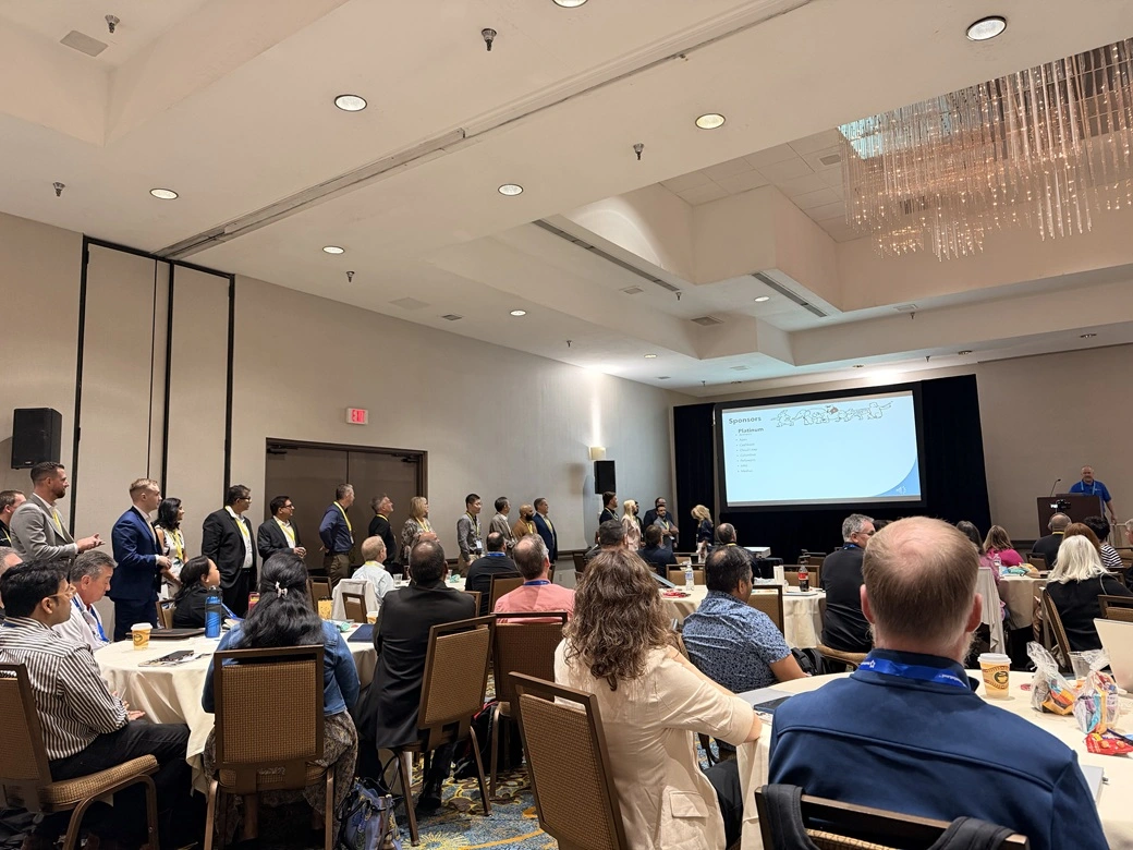Attendees at the 2026 M3UGA Spring Conference seated at round tables, viewing a Novacura presentation on a large screen at the front of a professional meeting room. A speaker stands at a podium while a group of participants stands along the wall observing.