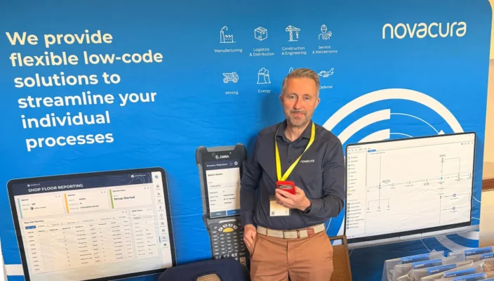 Man standing at a Novacura trade show booth, holding a smartphone and wearing a yellow lanyard, in front of a blue backdrop that reads “We provide flexible low-code solutions to streamline your individual processes.