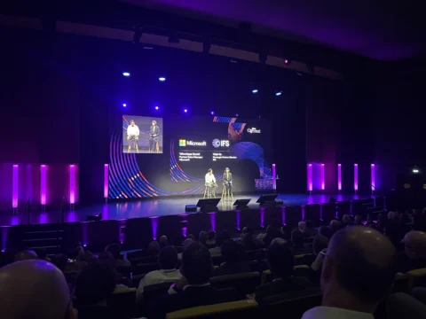 A conference presentation taking place on a large stage with purple lighting, where two speakers are seated and discussing in front of a large audience. The screen behind them displays IFS branding during an IFS Connect event.