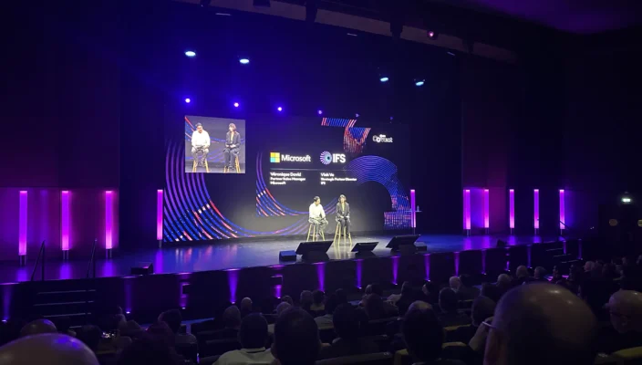 A conference presentation taking place on a large stage with purple lighting, where two speakers are seated and discussing in front of a large audience. The screen behind them displays IFS branding during an IFS Connect event.