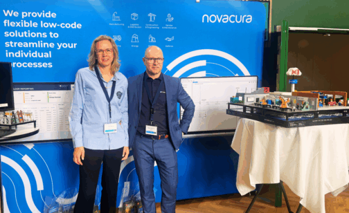 Two Novacura representatives standing at a company exhibition booth with a blue branded backdrop displaying the Novacura logo and icons for industries including Manufacturing, Logistics, Energy, and Defense.