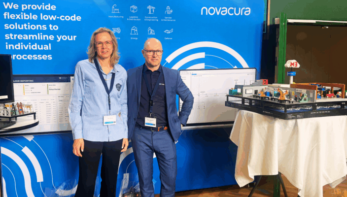 Two Novacura representatives standing at a company exhibition booth with a blue branded backdrop displaying the Novacura logo and icons for industries including Manufacturing, Logistics, Energy, and Defense.